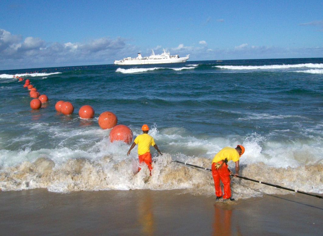 foto d&rsquo;operaris a la platja, cable que ve del mar, boies i vaixell al fons